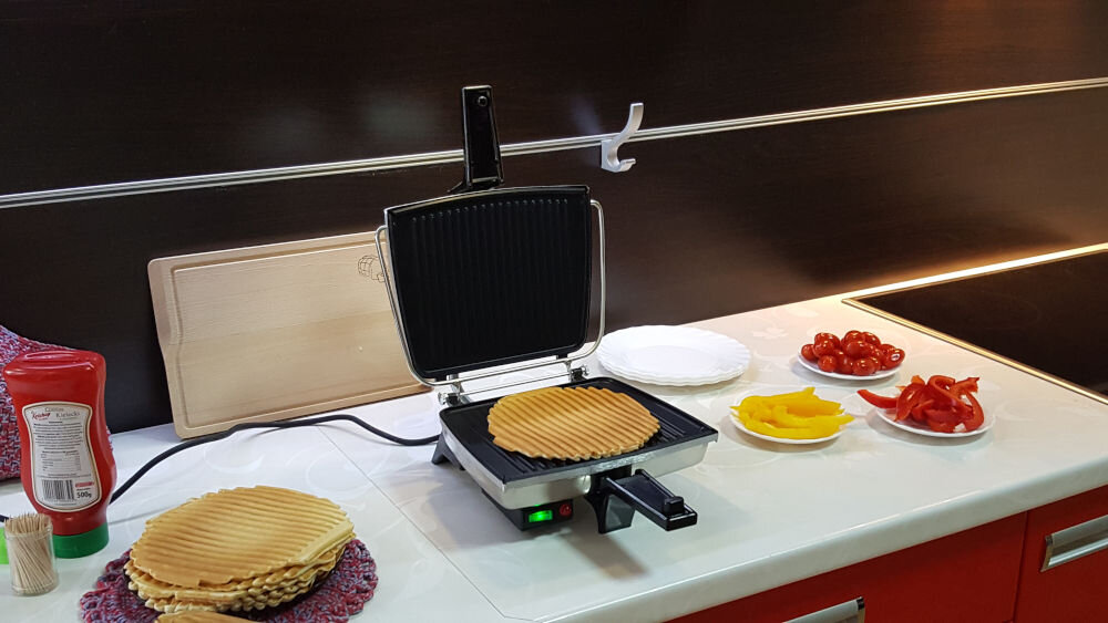 Opiekacz DEZAL Biały otwarty opiekacz czarny środek wewnątrz upieczony naleśnik urządzenie stoi na blacie wokół na talerzach składniki wszechstronny główne przeznaczenie pieczenie mięsa wypiekanie gofrów ciasto biszkoptowe wytrawne opiekanie kanapek pity tortille do codziennego użytku przygotowywanie różnorodnych potraw niezastąpiony w kuchni zdrowa zróżnicowana dieta