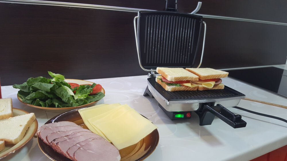 Opiekacz DEZAL Biały opiekacz stoi na stole świeci się włącznik wewnątrz urządzenia kanapki obok urządzenia znajdują się talerze z rozłożonymi składnikami system kontroli temperatury odpowiednie warunki do pieczenia płyty grzewcze stop aluminium pokryte powłoką greblonową zapobieganie przywieraniu ułatwienie przygotowanie łatwiejsze czyszczenie lampki kontrolne stan urządzenia zielona połączenie do sieci czerwona rozpoczęcie procesu grzania wbudowany ogranicznik temperatury bezpieczeństwo efektywność 