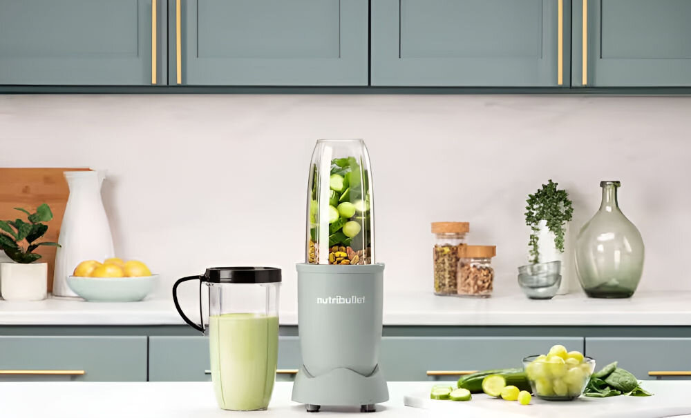 Na blacie kuchennym stoi blender personalny z logo \'nutribullet\', wypełniony kawałkami owoców i orzechów. Obok znajduje się kubek z zielonym napojem, a w tle widoczne są szafki kuchenne i dekoracje.