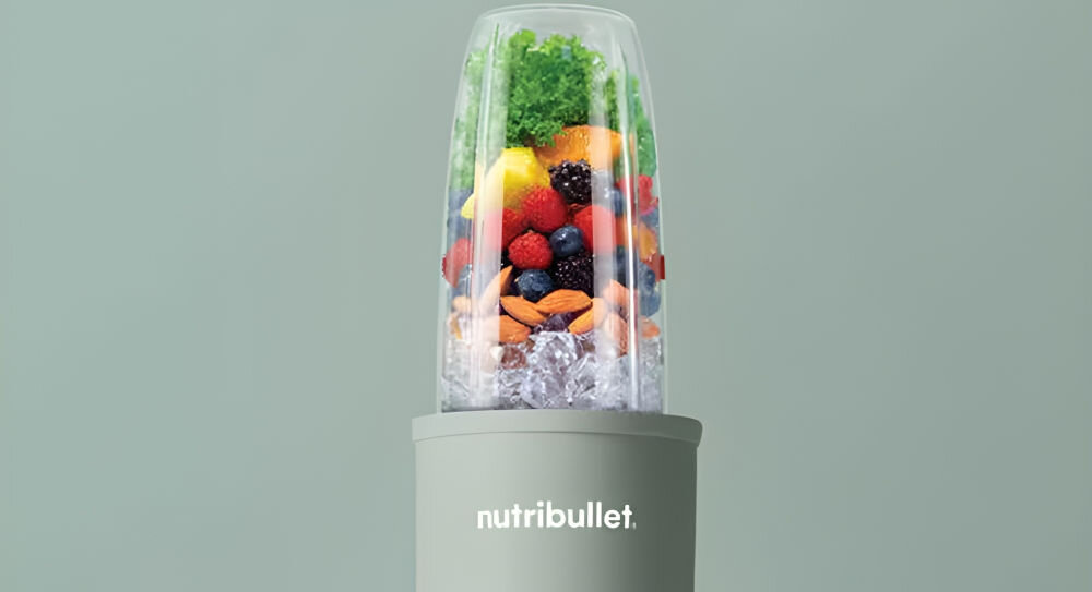 Blender z przezroczystym pojemnikiem wypełnionym owocami, warzywami i lodem, na obudowie widoczne logo \'nutribullet\'.