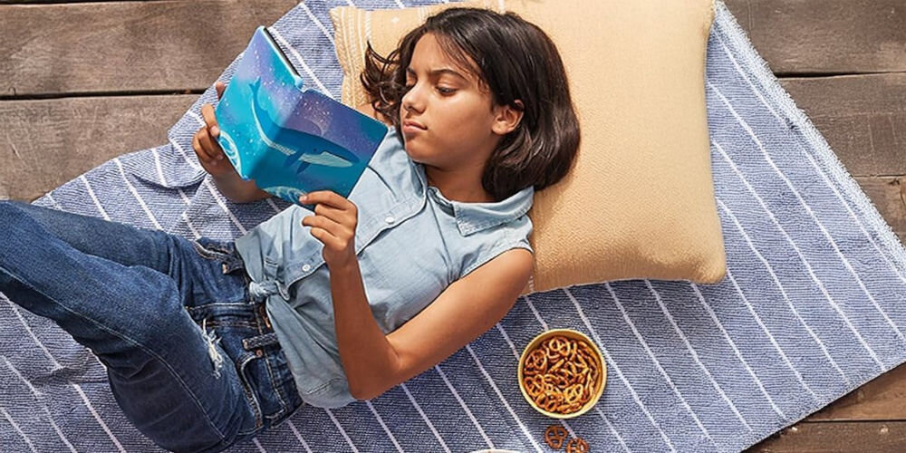Czytnik e-booków AMAZON Kindle Kids Space Whale 6 cali, Podświetlany ekran, Wi-Fi, Bez reklam Dziewczynka leży na kocu z poduszką, czyta Kindle Kids w etui z motywem wieloryba, obok miska z precelkami kontrola nad treściami Parent Dashboard brak reklam wsparcie rozwoju zdrowe nawyki czytelnicze