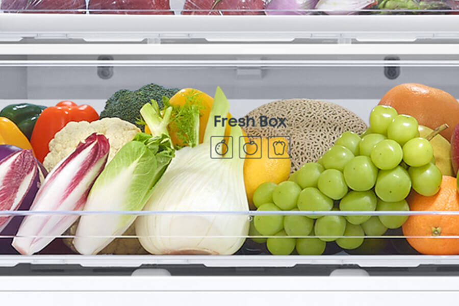 Wnętrze lodówki z szufladą oznaczoną napisem \'Fresh Box\', wypełnioną różnorodnymi warzywami i owocami, takimi jak winogrona, sałata, brokuły i pomarańcze.
