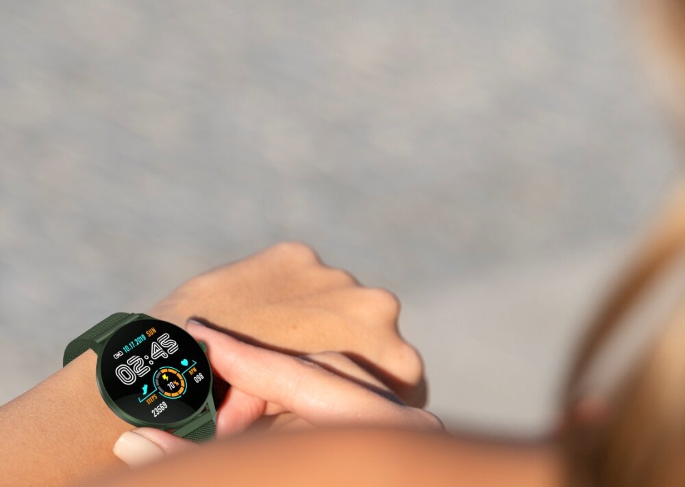 Osoba nosi smartwatch z okrągłym wyświetlaczem, na którym widoczna jest godzina 05:45 oraz data. Tło jest rozmyte, a pasek zegarka ma ciemny kolor.