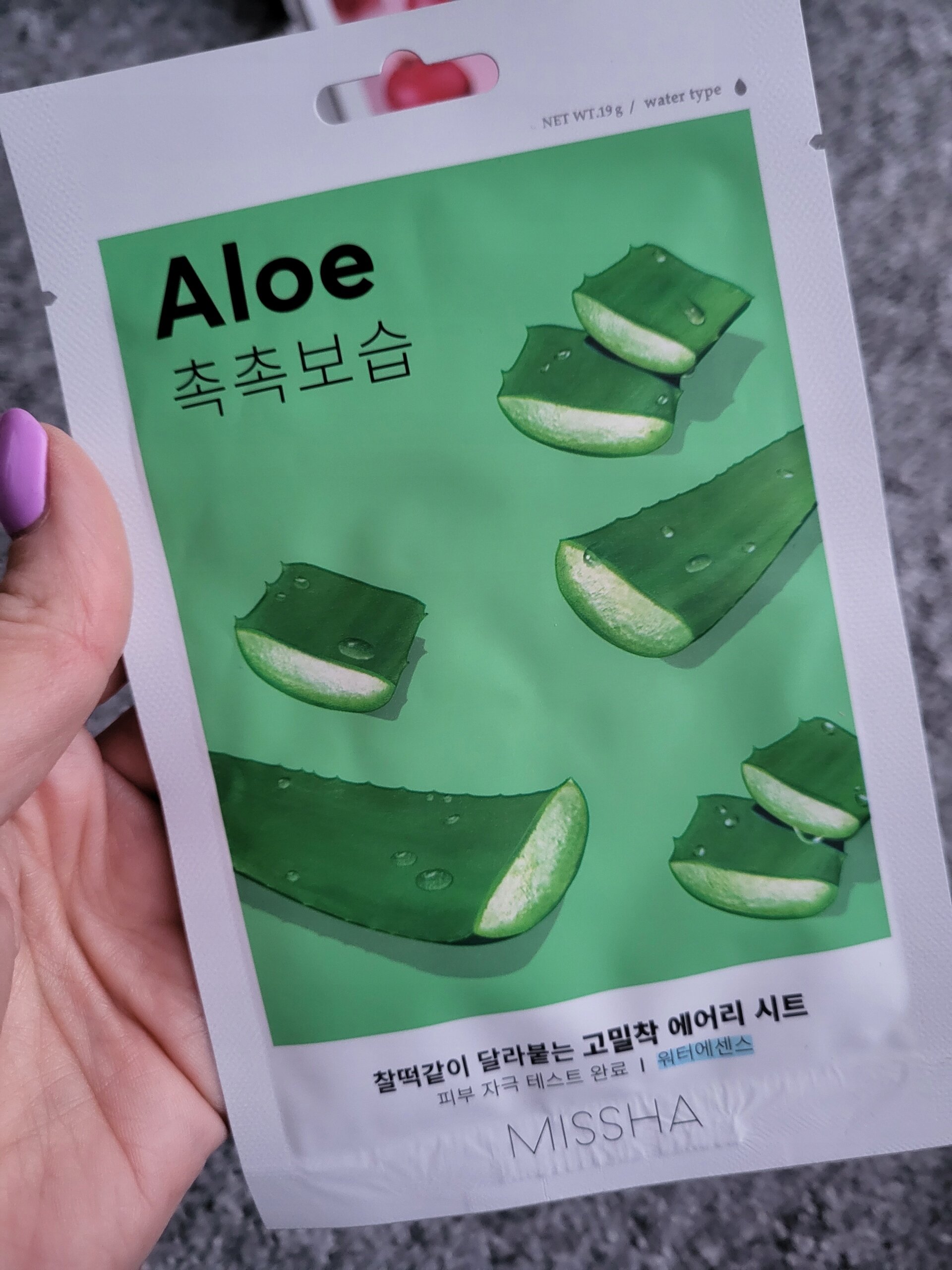Opakowanie maseczki w płachcie z aloesem, z widocznymi kawałkami aloesu na zielonym tle. Na opakowaniu znajduje się tekst: \'Aloe\', \'MISSHA\'.