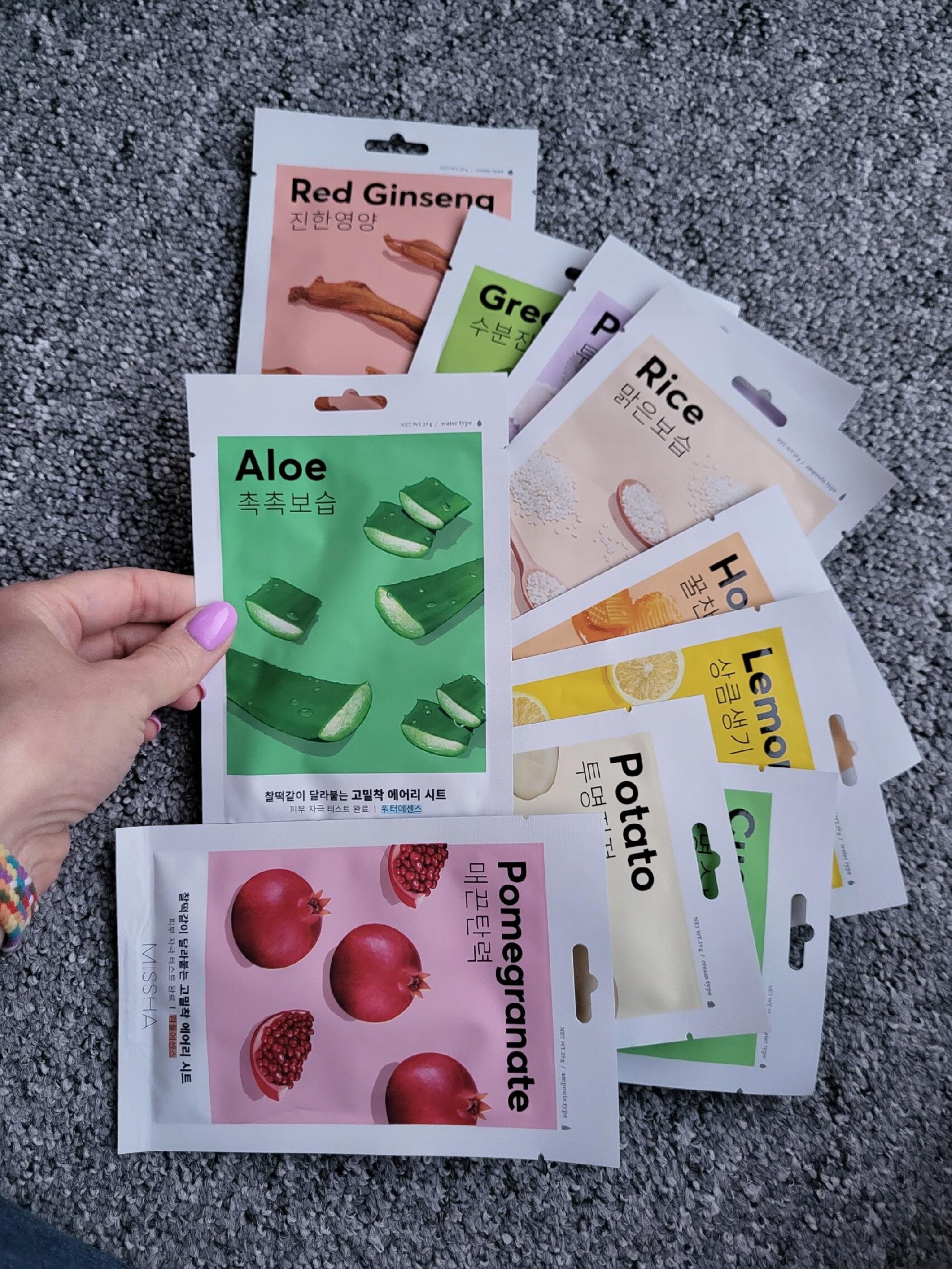 Na obrazie widocznych jest kilka opakowań maseczek w płachcie, w tym \'Aloe\' z grafiką aloesu oraz \'Pomegranate\' z grafiką granatu. Opakowania mają różne kolory i napisy w języku angielskim oraz koreańskim.