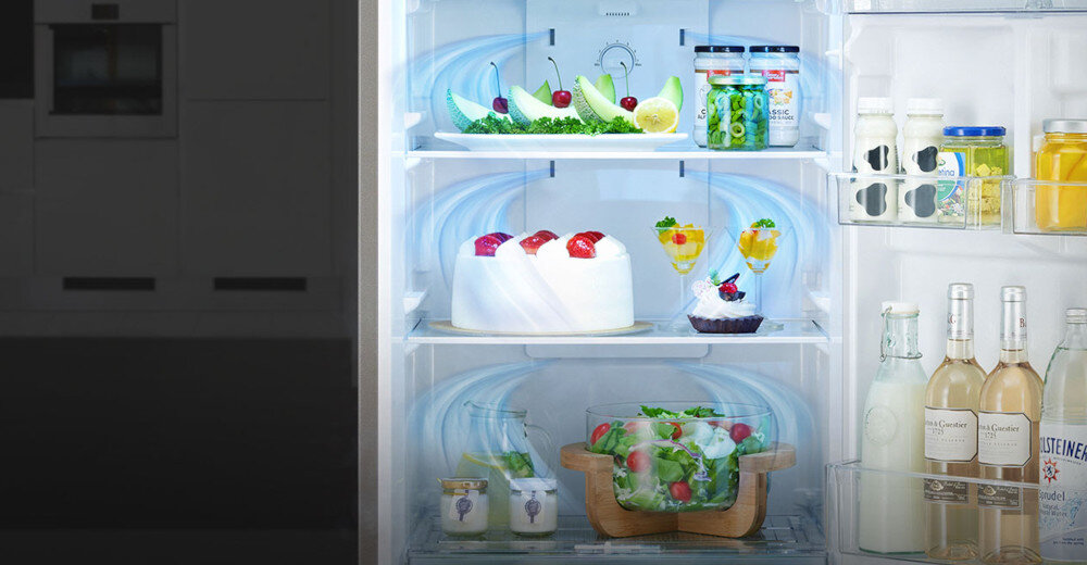 Wnętrze lodówki z różnorodnymi produktami spożywczymi, w tym ciastem, sałatką, napojami i jogurtami. Widoczne są również butelki z napojami na drzwiach lodówki.
