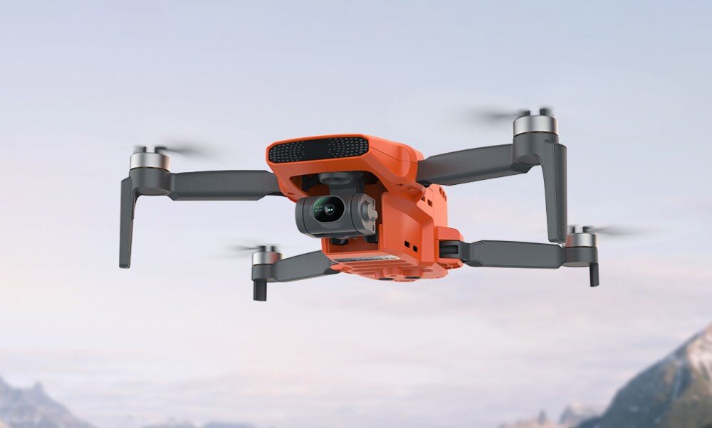 Na obrazie widoczny jest pomarańczowy dron z czterema śmigłami, unoszący się w powietrzu na tle nieba i gór. Dron ma zamontowaną kamerę z przodu.