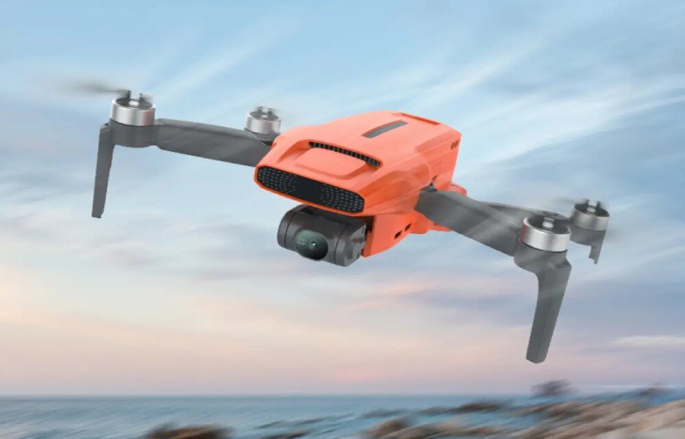 Pomarańczowy dron z czterema śmigłami unosi się w powietrzu na tle nieba i morza. Dron posiada kamerę zamontowaną z przodu.