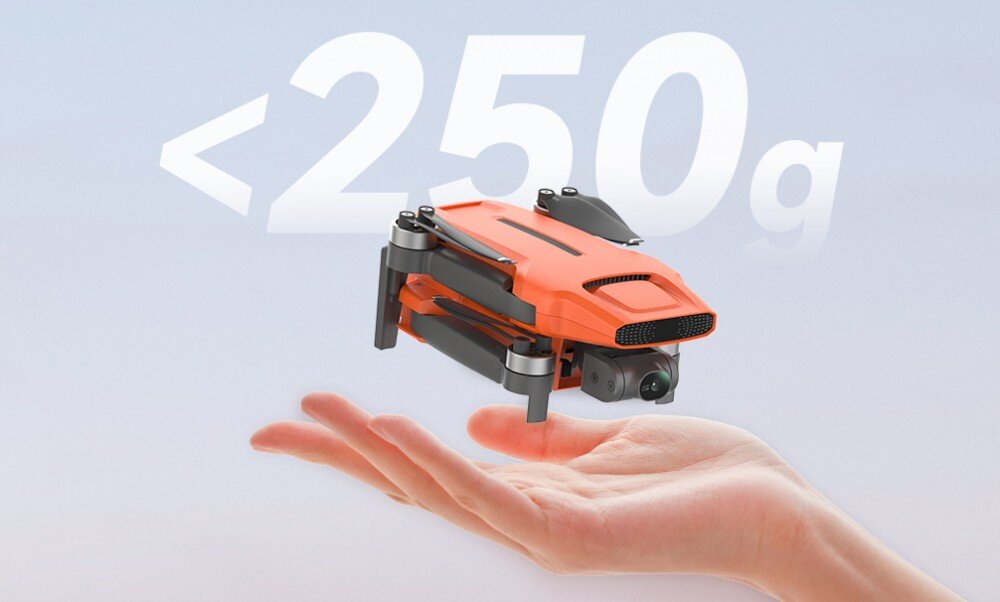 Pomarańczowy dron unosi się nad otwartą dłonią na tle nieba. Widoczny jest napis \'<250g\'.
