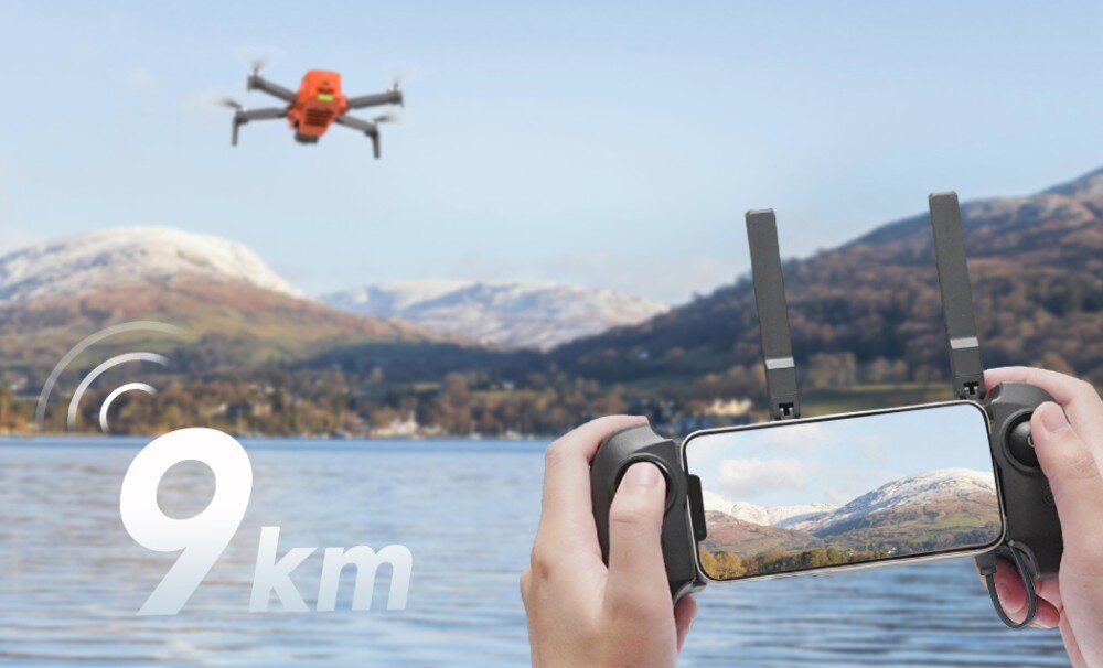 Osoba trzyma kontroler z ekranem, na którym widać krajobraz górski, a w tle unosi się pomarańczowy dron. Na obrazie widnieje napis \'9 km\'.