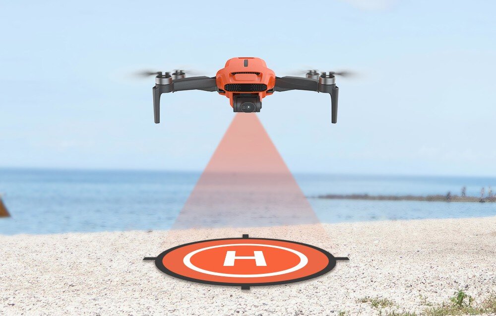 Pomarańczowy dron unosi się nad plażą, emitując czerwony promień skierowany na okrągłą matę z literą \'H\'. W tle widać morze i niebo.