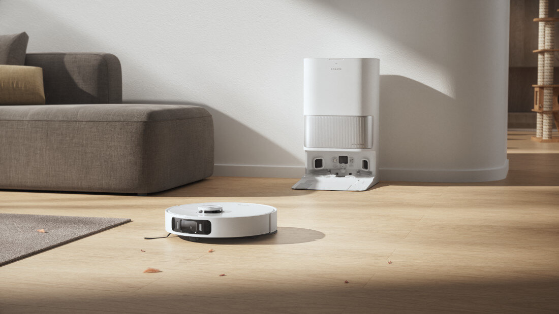 Na drewnianej podłodze znajduje się biały robot sprzątający, a w tle widoczna jest stacja dokująca przy ścianie. Obok znajduje się szara kanapa, a na podłodze leżą pojedyncze liście.