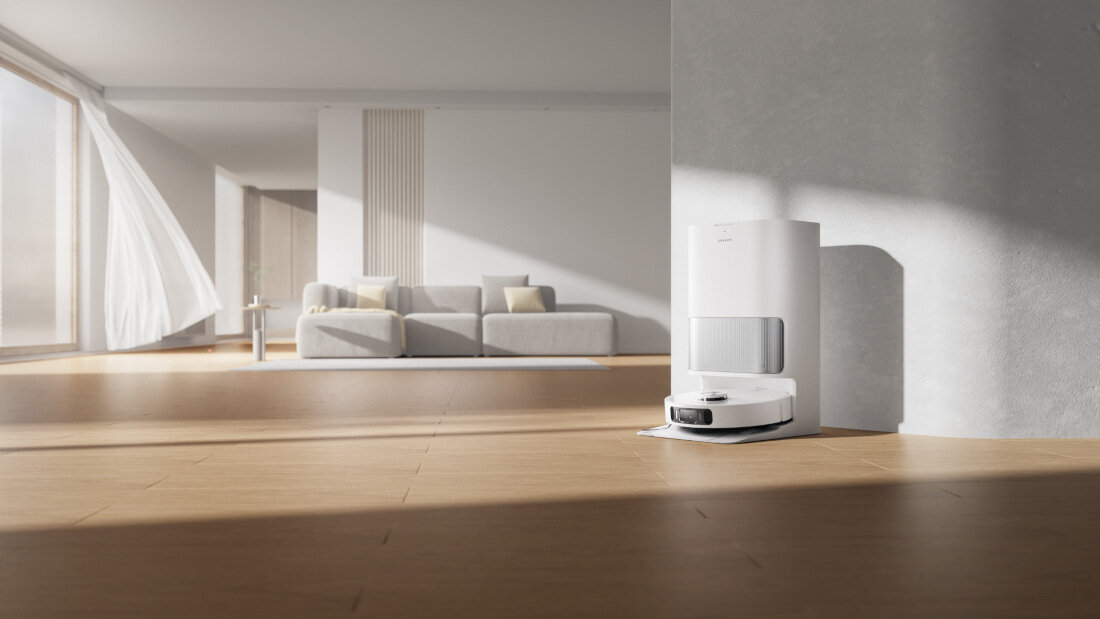 Na drewnianej podłodze w jasnym pomieszczeniu znajduje się biały robot sprzątający z bazą ładującą. W tle widać sofę z poduszkami oraz duże okno z zasłoną.