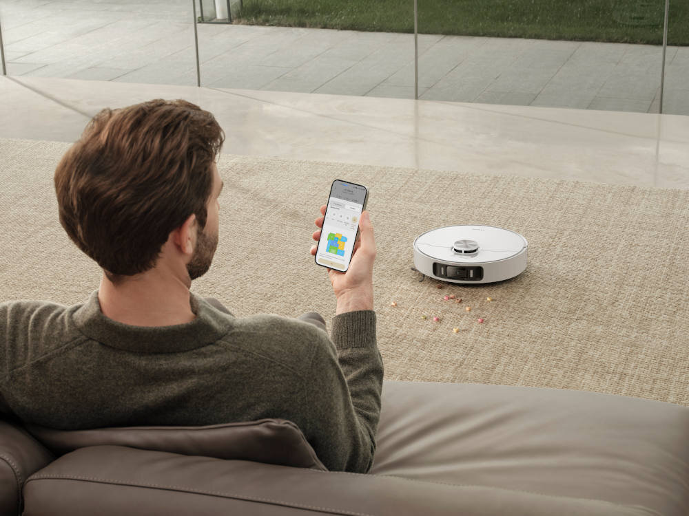 Osoba siedzi na kanapie, trzymając w ręku smartfon z aplikacją pokazującą mapę. Na dywanie obok znajduje się biały robot sprzątający, wokół którego leżą rozsypane drobinki.