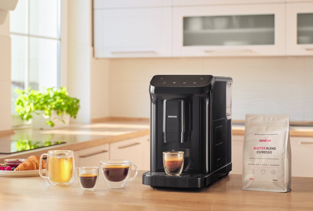 Czarny ekspres do kawy marki SENCOR stoi na blacie kuchennym obok trzech filiżanek z różnymi napojami oraz opakowania kawy z napisem \'MASTER BLEND ESPRESSO\'.