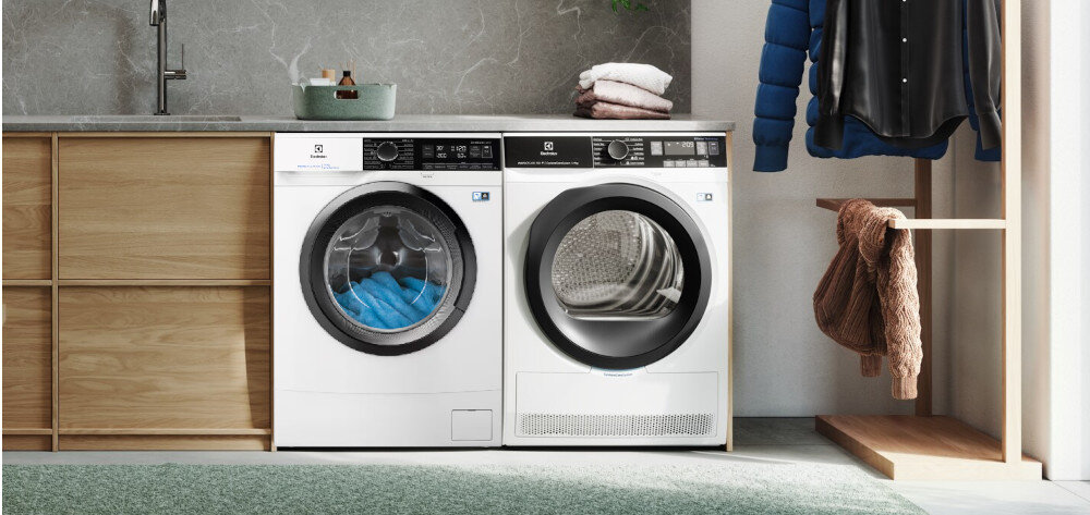 Na obrazie widoczne są dwie białe pralki marki Electrolux ustawione obok siebie w nowoczesnej łazience. Na pralce po lewej stronie znajduje się napis \'ELECTROLUX\', a na pralce po prawej stronie widoczny jest bęben suszarki.
