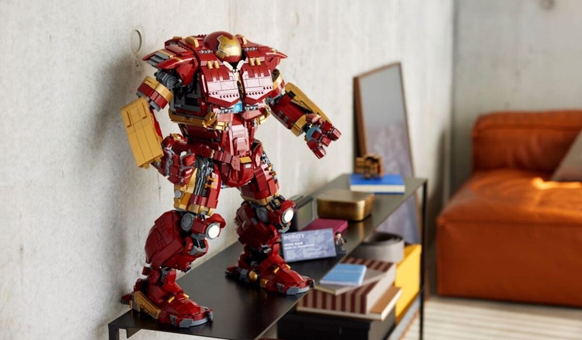 Na półce stoi duży model z klocków w kształcie czerwono-złotego robota. W tle widoczna jest pomarańczowa sofa i kilka książek na półce.