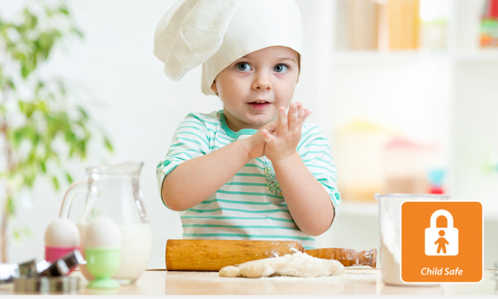 Dziecko w czapce kucharskiej stoi przy stole z wałkiem do ciasta i składnikami do pieczenia. W prawym dolnym rogu znajduje się pomarańczowa ikona z napisem \'Child Safe\' i symbolem kłódki.