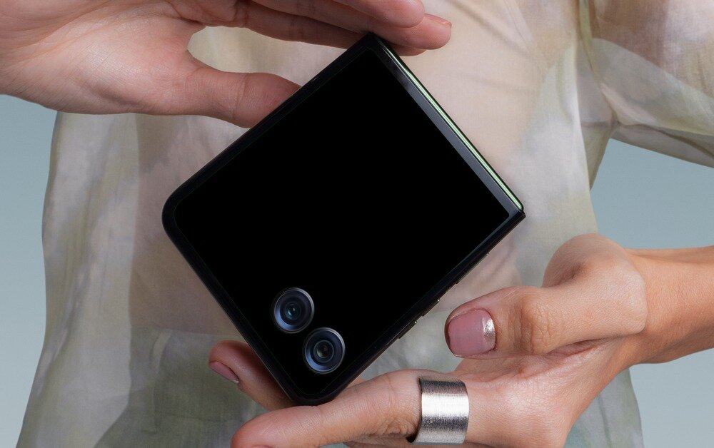 Osoba trzyma złożony smartfon z widocznymi dwoma obiektywami aparatu na czarnym tle obudowy.