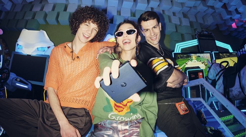 Trzy osoby pozują do zdjęcia w jasnym pomieszczeniu z neonowymi światłami, jedna z nich trzyma smartfon z widocznym logo producenta. W tle znajdują się elementy przypominające sprzęt do gier.