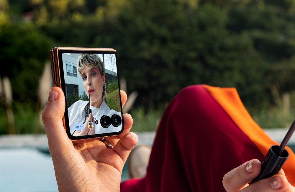 Osoba trzyma smartfon z dużym ekranem, na którym widać selfie kobiety z krótkimi włosami. W tle znajduje się rozmyty krajobraz z zielenią.