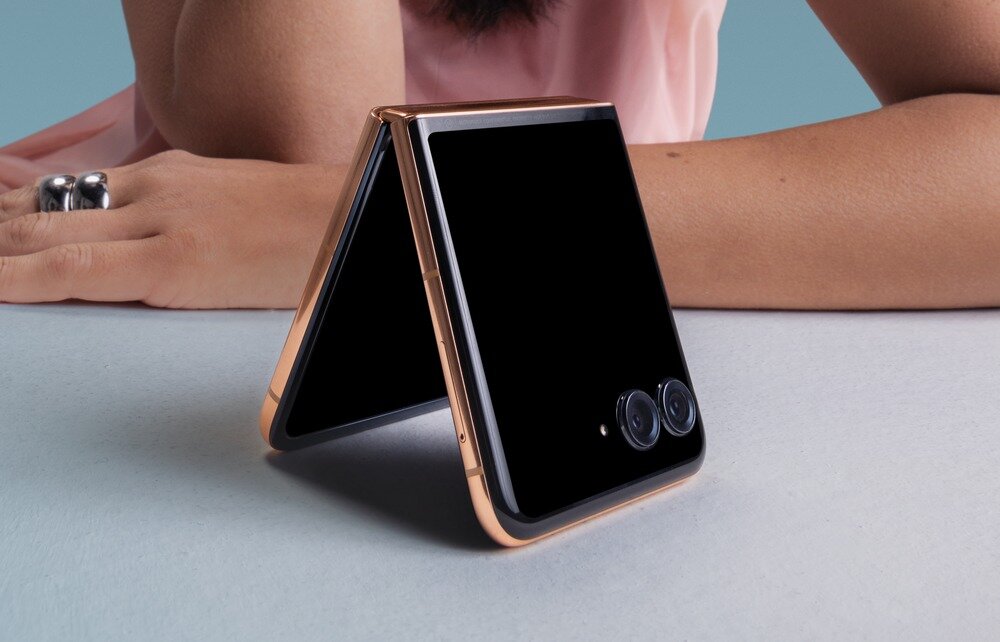 Na obrazie widoczny jest składany smartfon w kształcie litery A z dwoma obiektywami aparatu na zewnętrznej stronie. W tle znajduje się osoba opierająca się na rękach.