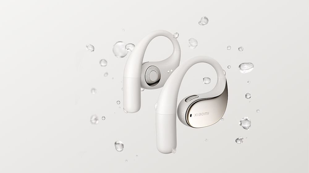 Na obrazie widoczne są beżowe słuchawki douszne z logo \'XIAOMI\' na jednej z nich. Wokół słuchawek unoszą się krople wody na jasnym tle.