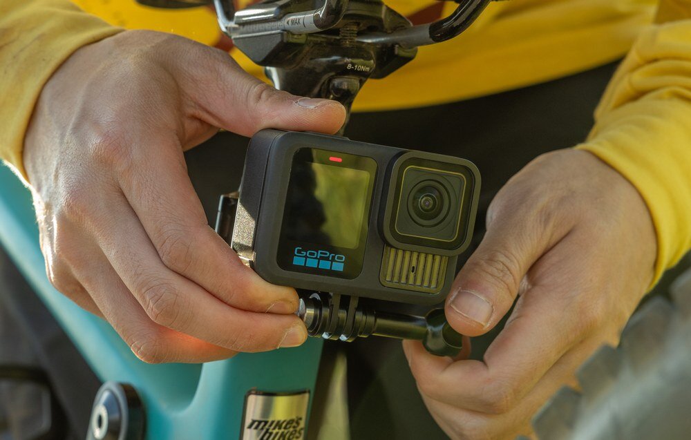 Osoba trzyma kamerę sportową z logo \'GoPro\' zamontowaną na rowerze. Kamera jest czarna i ma widoczny obiektyw oraz ekran.