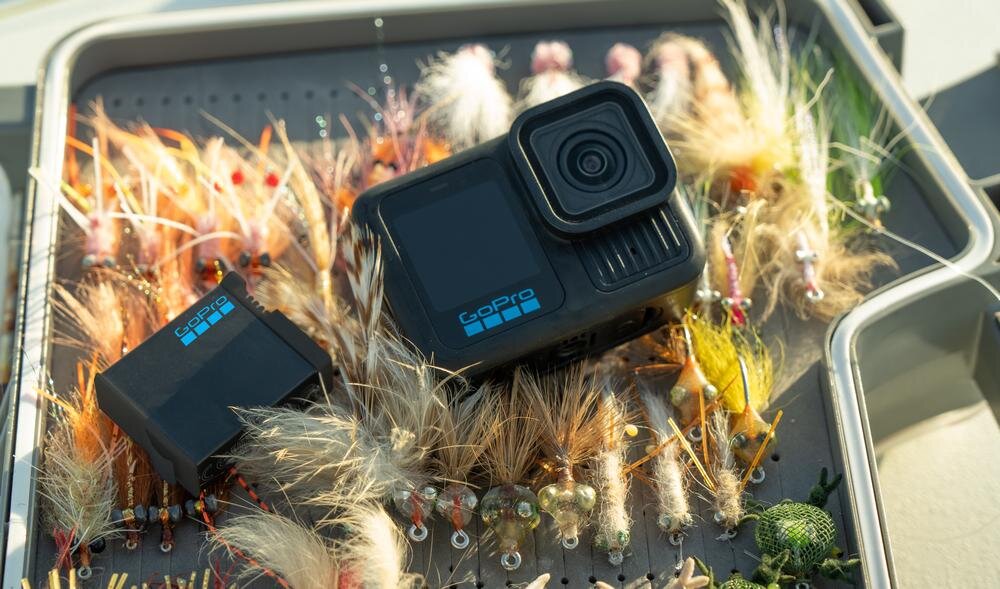 Kamera sportowa z logo GoPro leży na pudełku z różnorodnymi sztucznymi muchami wędkarskimi. Obok kamery znajduje się akumulator również z logo GoPro.