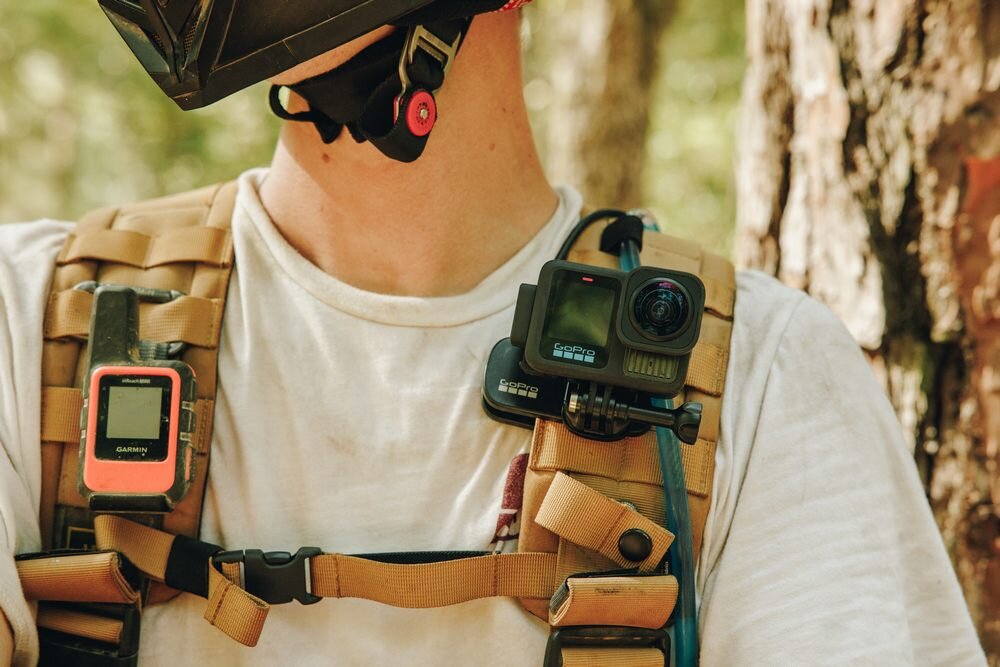Osoba w białej koszulce nosi na piersi zamocowaną kamerę sportową z napisem \'GOPRO HERO 13 BLACK\'. Obok kamery znajduje się urządzenie z logo \'Garmin\'.