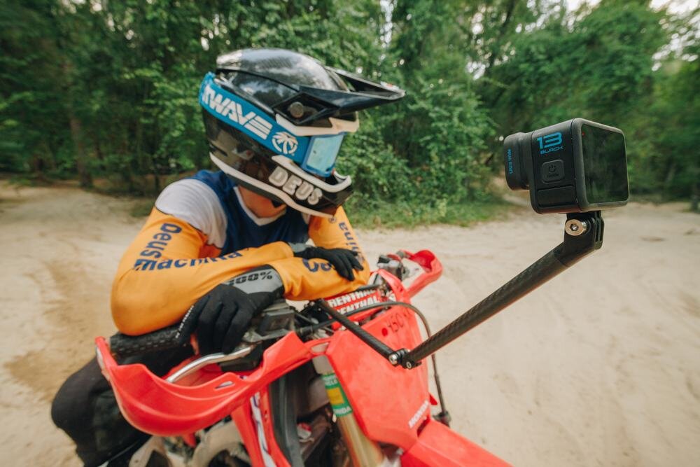 Osoba w kasku siedzi na czerwonym motocyklu terenowym, a na wysięgniku zamontowana jest kamera sportowa z napisem \'13 BLACK\'. W tle widoczne są drzewa i piaszczysta droga.