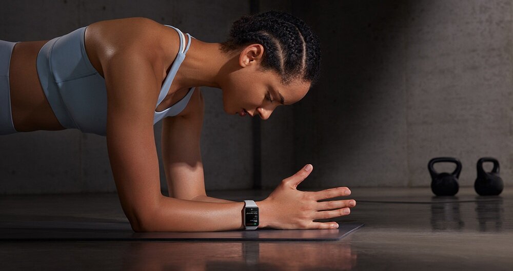 Osoba w sportowym stroju wykonuje ćwiczenie na macie, nosząc czarny smartband na nadgarstku. W tle widoczne są dwa kettlebelle.