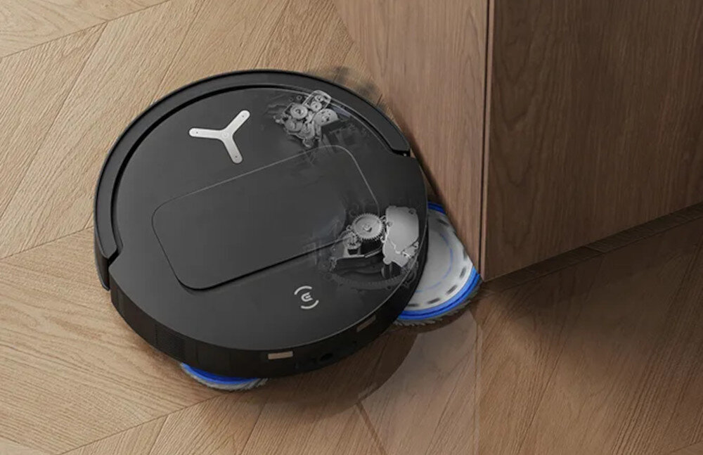 Czarny robot sprzątający z widocznym logo na górze, poruszający się przy drewnianej szafce na podłodze z jasnego drewna. Na robocie widoczne są przezroczyste elementy mechanizmu.