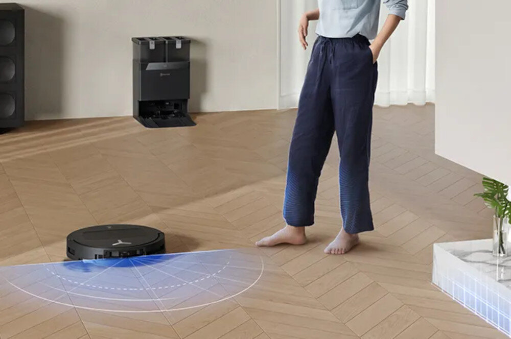 Na drewnianej podłodze znajduje się czarny robot sprzątający z niebieskim światłem emitowanym z przodu. W tle widać stację dokującą oraz osobę stojącą obok robota.