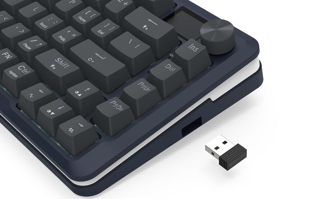 Na obrazie widoczna jest klawiatura z ciemnymi klawiszami i pokrętłem w rogu. Obok klawiatury znajduje się mały adapter USB.