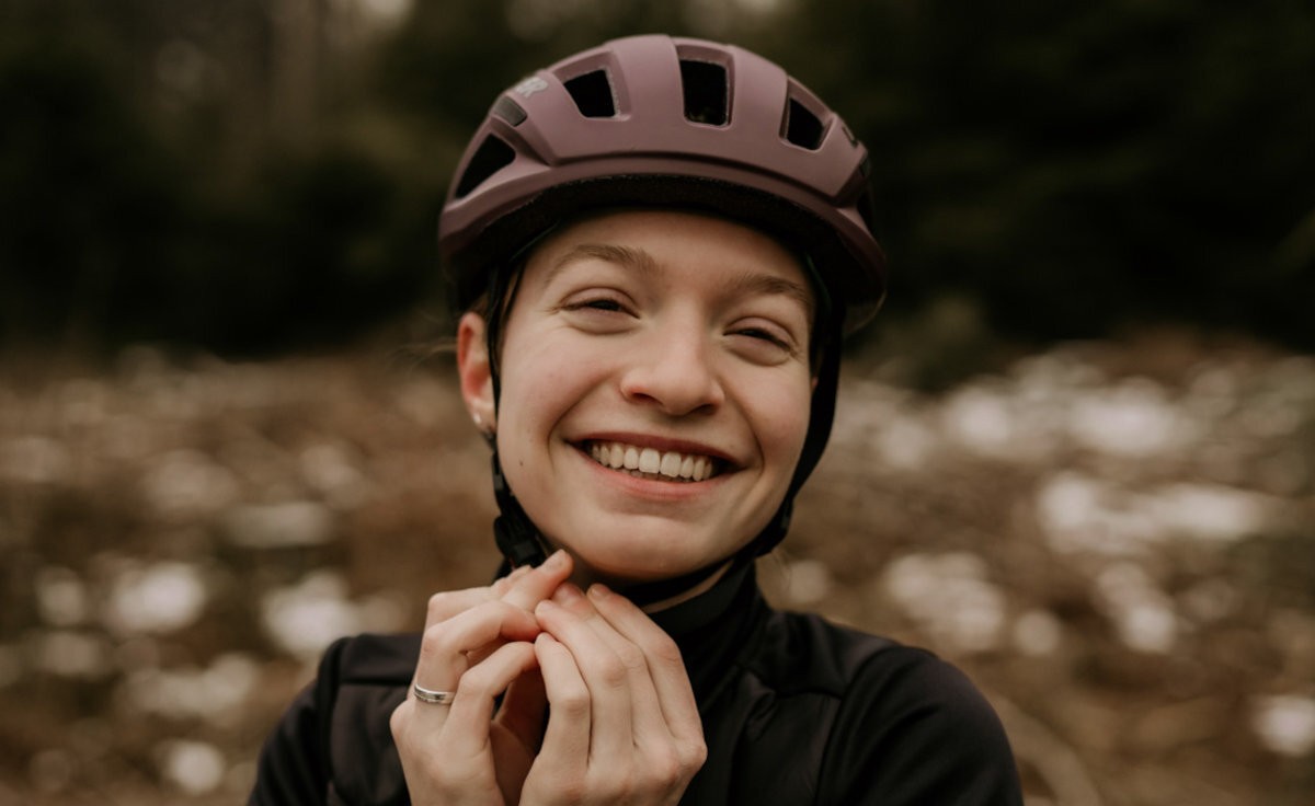 Osoba w kasku rowerowym stoi na tle rozmytego krajobrazu z drzewami i śniegiem na ziemi.