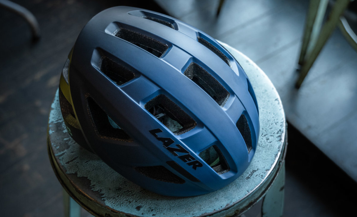 Na obrazie widoczny jest niebieski kask rowerowy z dużymi otworami wentylacyjnymi, leżący na okrągłym, metalowym stołku. Na kasku znajduje się napis \'LAZER\'.