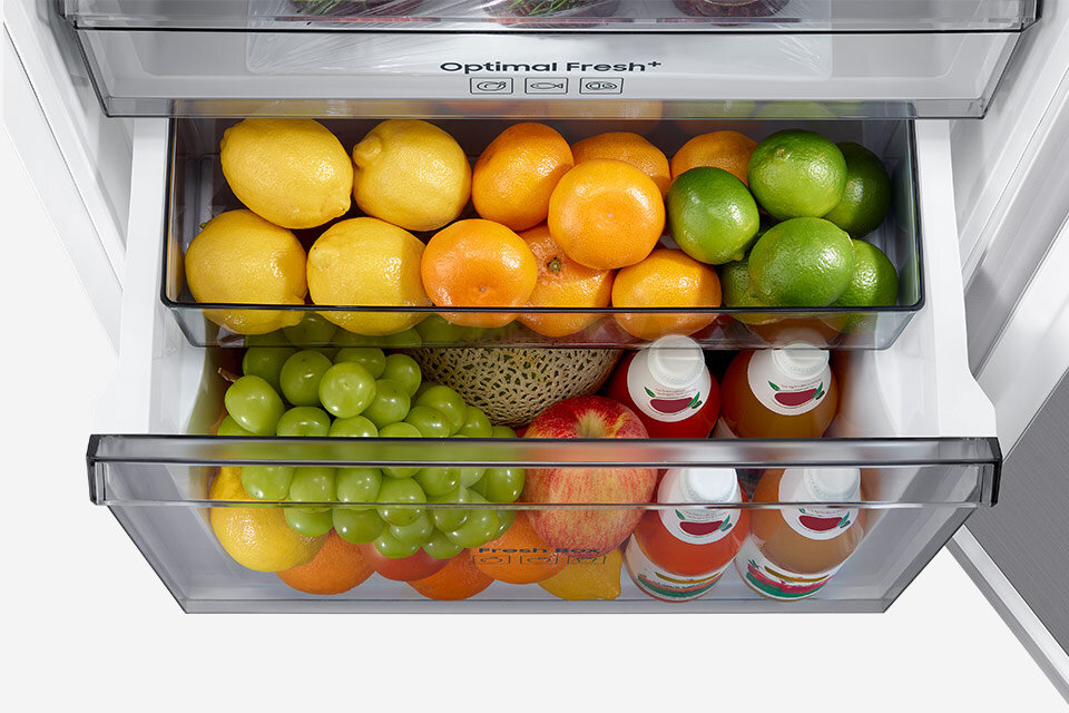 W lodówce znajdują się dwie szuflady wypełnione owocami, takimi jak cytryny, pomarańcze, limonki, winogrona, melon i jabłka. Na jednej z szuflad widoczny jest napis \'Optimal Fresh+\'.