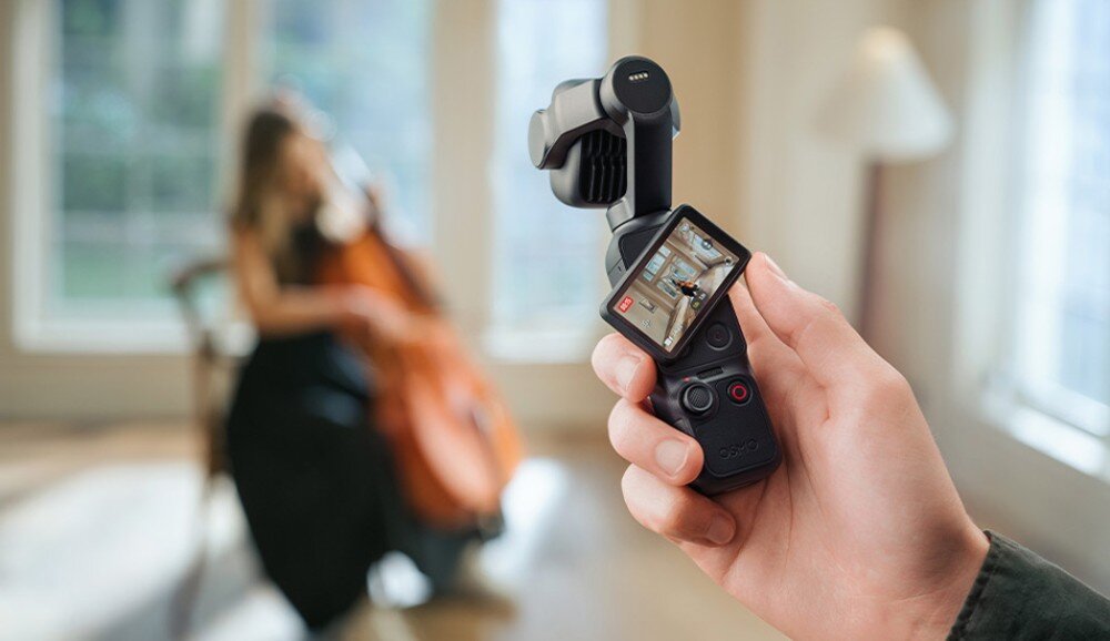 Kamera sportowa DJI Osmo Pocket 4P Dłoń trzyma kamerę z odchylonym ekranem pod kątem, na którym widać podgląd nagrywania kobiety z wiolonczelą