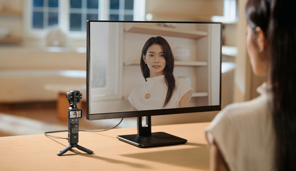 Kamera sportowa DJI Osmo Pocket 4P Kamera na małym statywie stoi na stole obok monitora wyświetlającego obraz kobiety w trakcie połączenia wideo