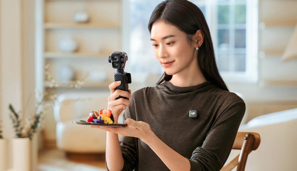 Kamera sportowa DJI Osmo Pocket 4P Kobieta siedząca przy stole trzyma w ręku małą kamerę na gimbalu i nagrywa talerz z owocowymi przekąskami