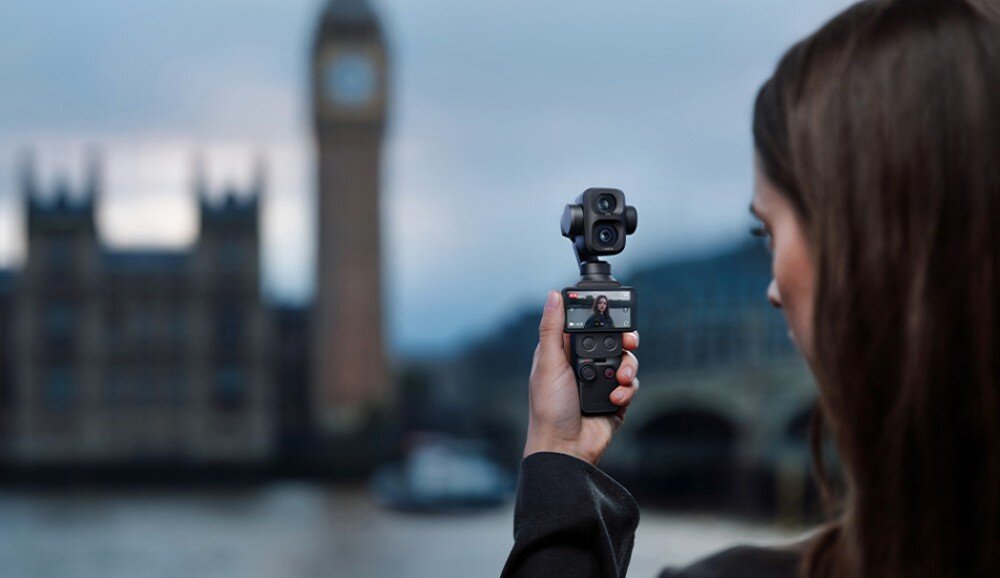 Kamera sportowa DJI Osmo Pocket 4P Osoba trzyma w dłoni małą kamerę z wyświetlaczem skierowaną w swoją stronę, nagrywając w tle słynną wieżę zegarową w Londynie