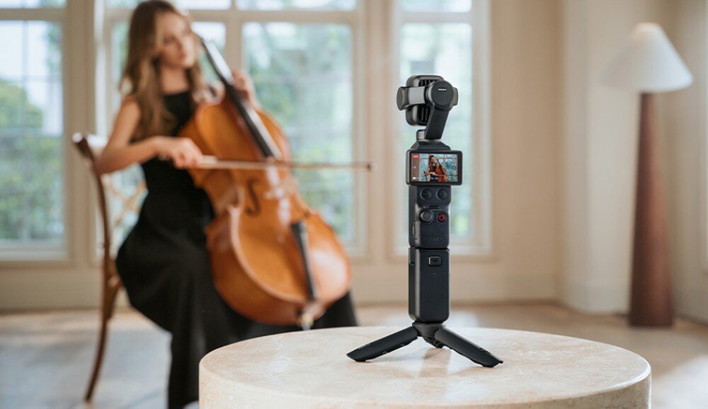 Kamera sportowa DJI Osmo Pocket 4P Kamera na małym statywie stoi na stole i nagrywa kobietę grającą na wiolonczeli w jasnym pomieszczeniu