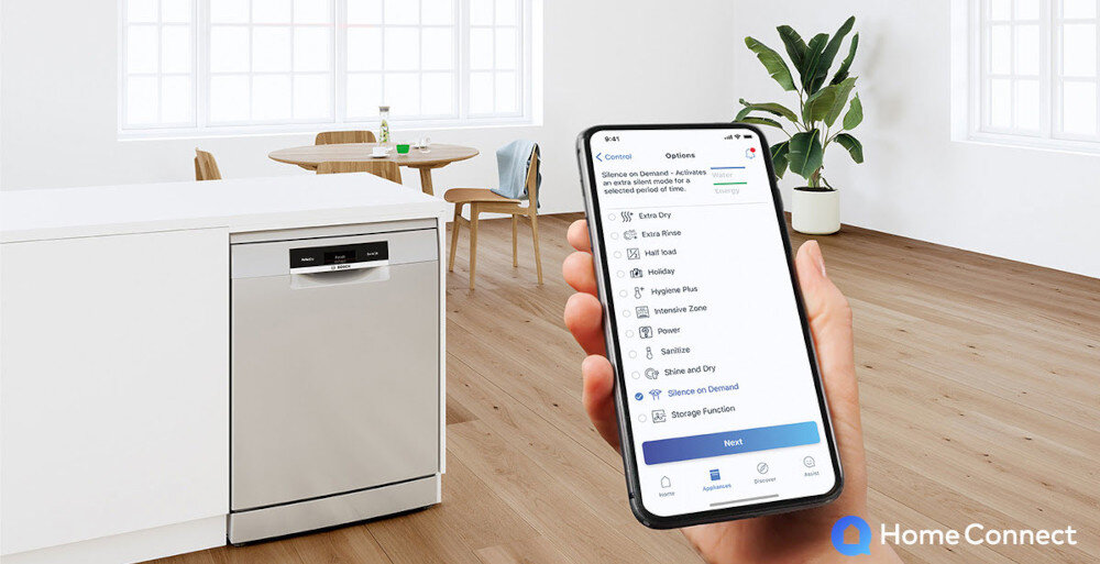 W jasnym wnętrzu kuchni znajduje się zmywarka o srebrnym froncie. Osoba trzyma smartfon z aplikacją Home Connect, na ekranie widoczne są opcje zmywania.