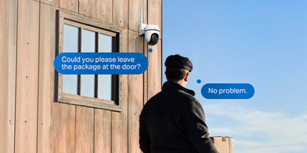 Na drewnianej ścianie budynku zamontowana jest kamera obrotowa. Obok niej widoczne są dymki z tekstem: \'Could you please leave the package at the door?\' oraz \'No problem.\'