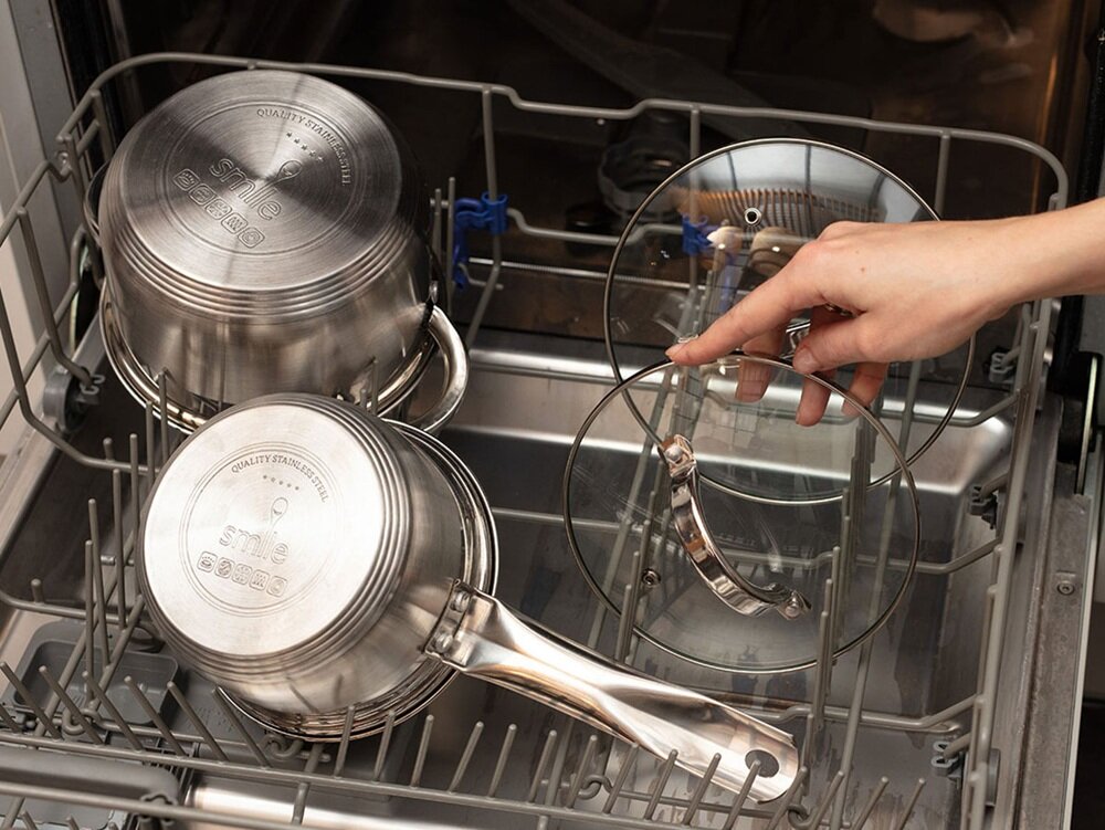 Wnętrze zmywarki z widocznymi metalowymi garnkami i szklanymi pokrywkami. Na garnkach widoczny jest napis \'QUALITY STAINLESS STEEL smile MGK-10\'.