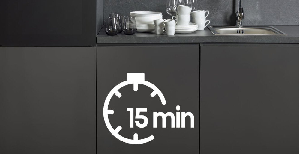 Na obrazie widoczna jest kuchnia z ciemnymi szafkami, na których znajduje się stos naczyń obok zlewu. Na froncie szafki widnieje symbol zegara z napisem \'15 min\'.