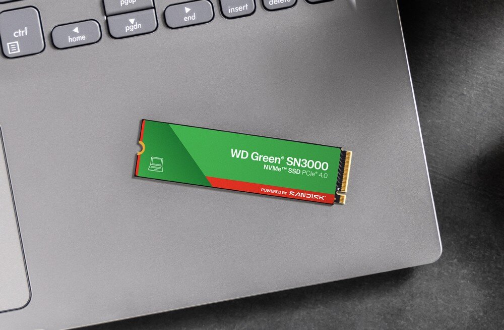 Na szarej powierzchni laptopa leży dysk SSD z napisem \'WD Green SN3000 NVMe SSD PCIe 4.0\' oraz logo \'SANDISK\'.