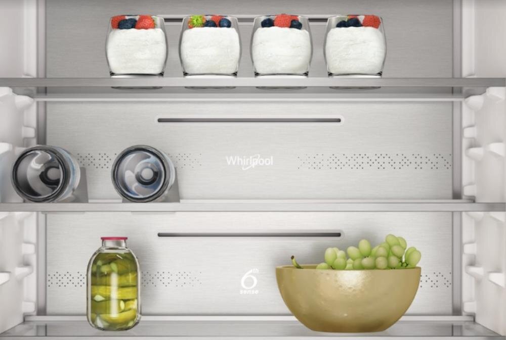 Wnętrze lodówki z trzema półkami, na których znajdują się jogurty z owocami, butelki, słoik z marynatami i miska z winogronami. Widoczne logo \'Whirlpool\' oraz symbol \'6th Sense\'.