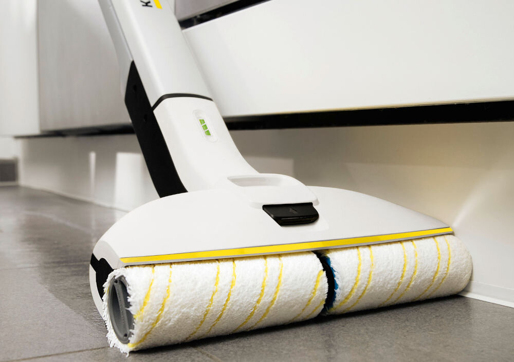 Na obrazie widoczny jest biały mop elektryczny z żółtymi akcentami, czyszczący podłogę. Na uchwycie znajduje się logo KARCHER.